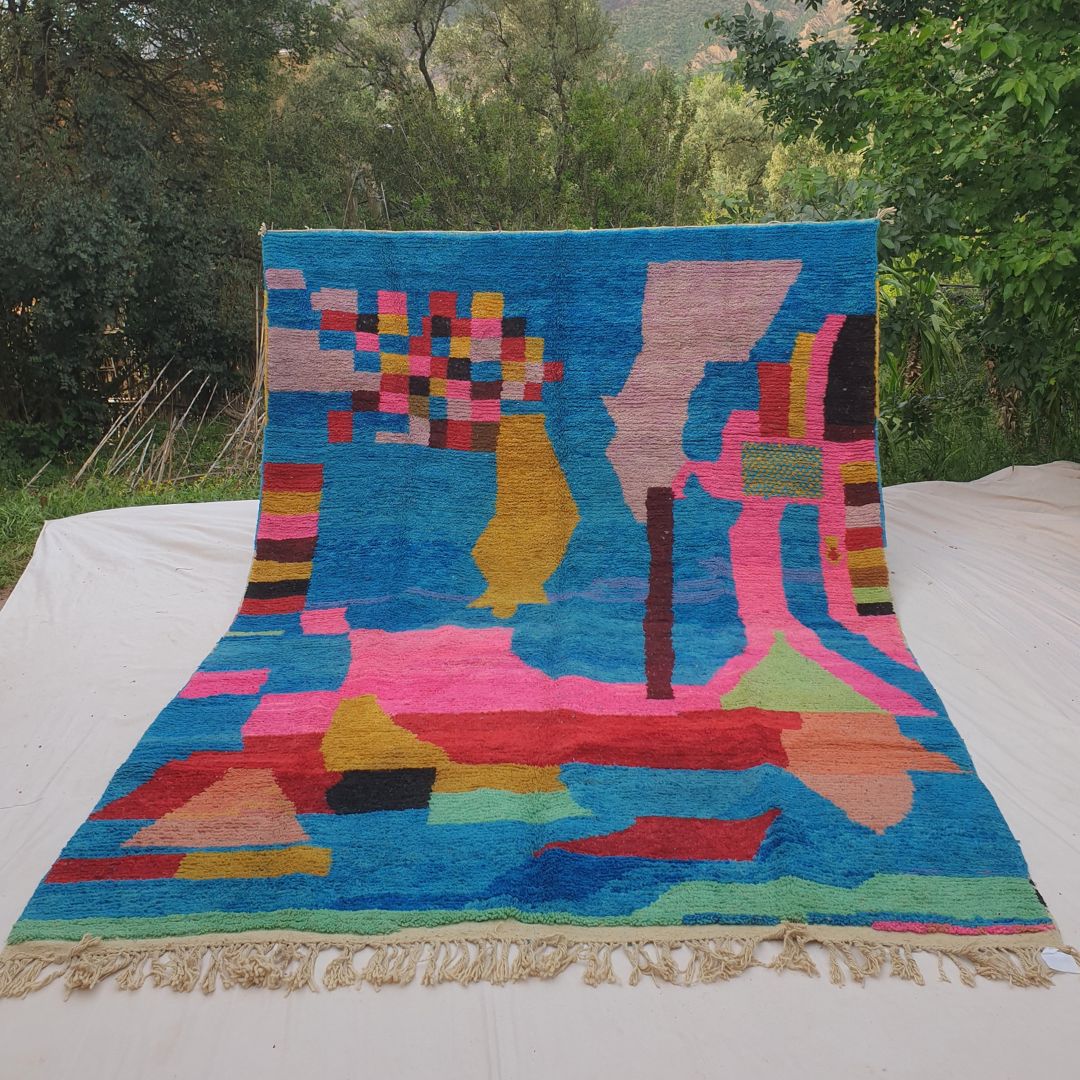 Person holding up a large electric blue Boujad Moroccan rug outdoors between trees, displaying its 257×360 cm size — a vivid cobalt blue field covered with bold abstract shapes in hot pink, red, mustard yellow, lime green, lavender and salmon, a striking contemporary Berber textile artwork