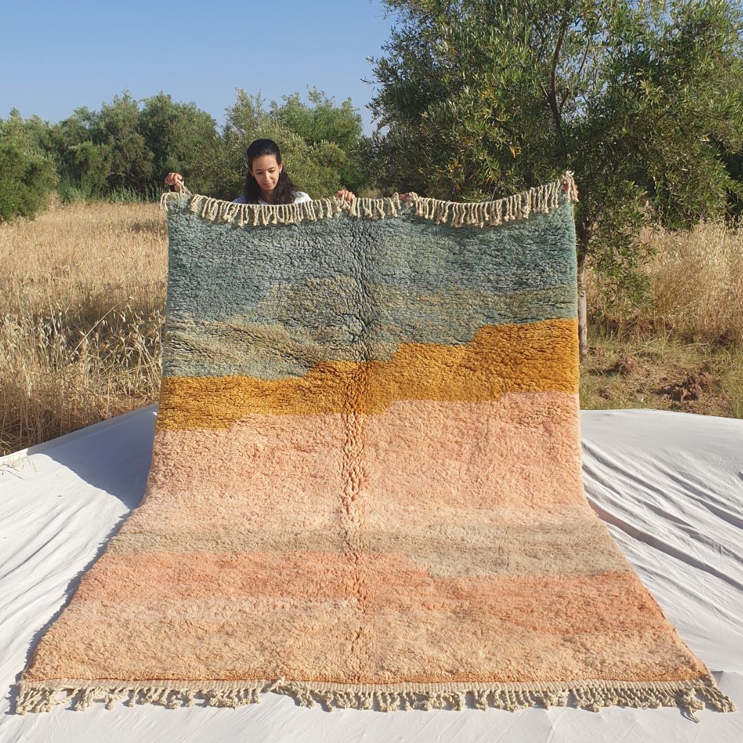 Person holding up a luxurious Beni Mrirt Moroccan rug outdoors in natural surroundings, showing its 201×304 cm size with a stunning painterly color-block composition — teal blue upper band, mustard yellow accent, and a warm salmon dusty rose dominant lower field creating a beautiful watercolor effect