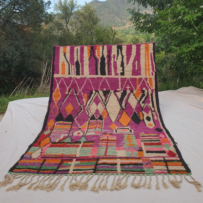 Person holding up a deep purple Boujad Moroccan rug outdoors between trees, displaying its 227×323 cm size — a dramatic violet-purple background with large white abstract silhouette shapes in the upper section, a row of orange and black diamonds in the middle, and a vivid multicolor stripe band at the bottom in green, orange, pink and white