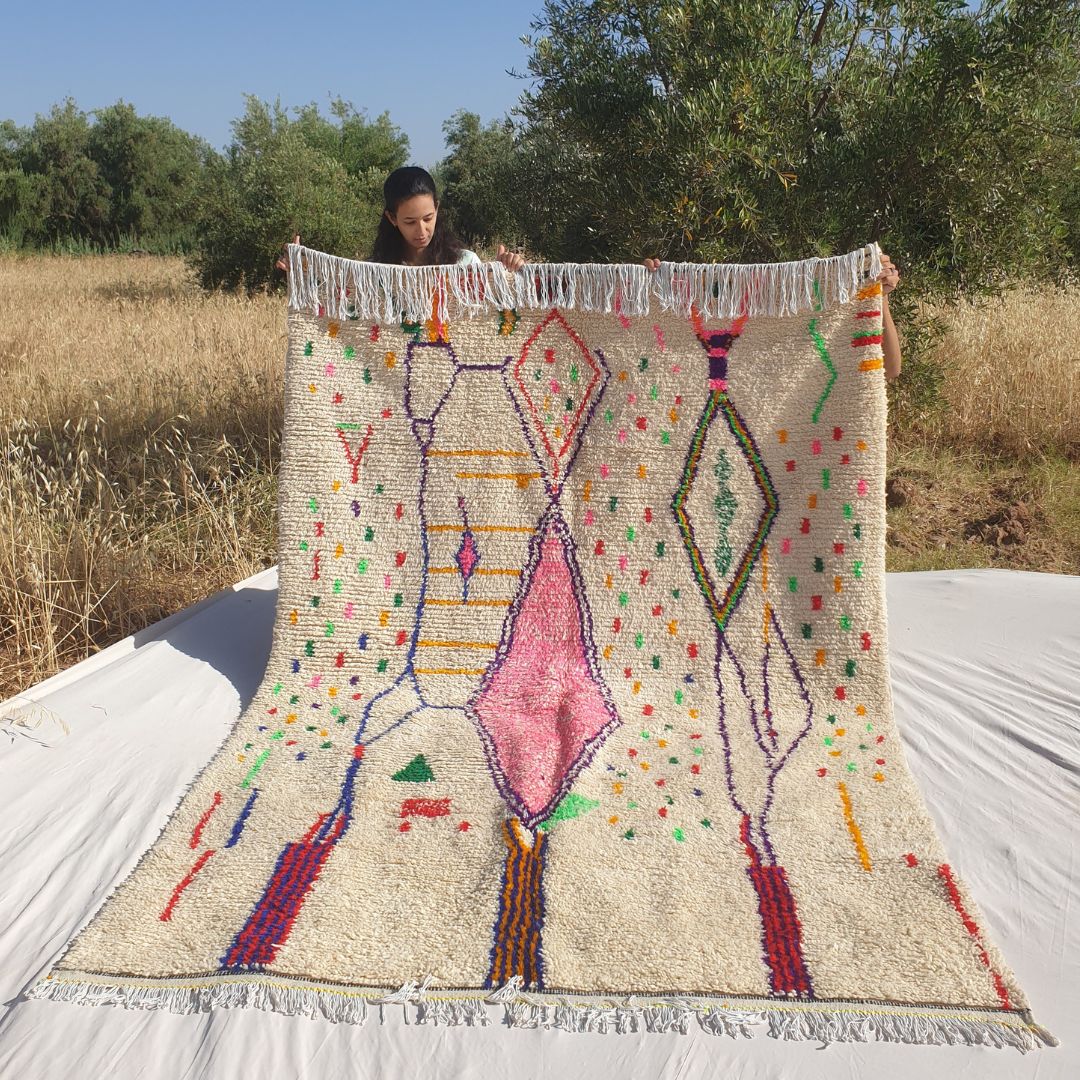 Person holding up a white Azilal Moroccan rug outdoors, showing its 195×282 cm size — a warm cream natural wool base scattered with joyful multicolor confetti dots in red, green, orange, yellow, blue and pink, with a large central rose-pink diamond, elongated purple diamond outlines, green outlined upper diamonds and bold navy and red vertical accent lines at the bottom