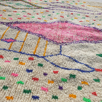 Close-up of a white Azilal Berber rug showing the natural cream wool pile densely scattered with tiny multicolor confetti dots and marks in red, green, orange, yellow and blue — the central rose-pink diamond motif and purple outlined elongated diamonds visible, all handwoven with extraordinary detail and Amazigh symbolism