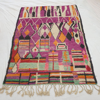 Authentic Boujad Moroccan rug 227×323 cm laid flat — deep violet purple background with large white abstract silhouette shapes in the upper zone, orange and black diamond row in the middle, and vivid multicolor stripes at the bottom, handmade in Morocco by Berber women