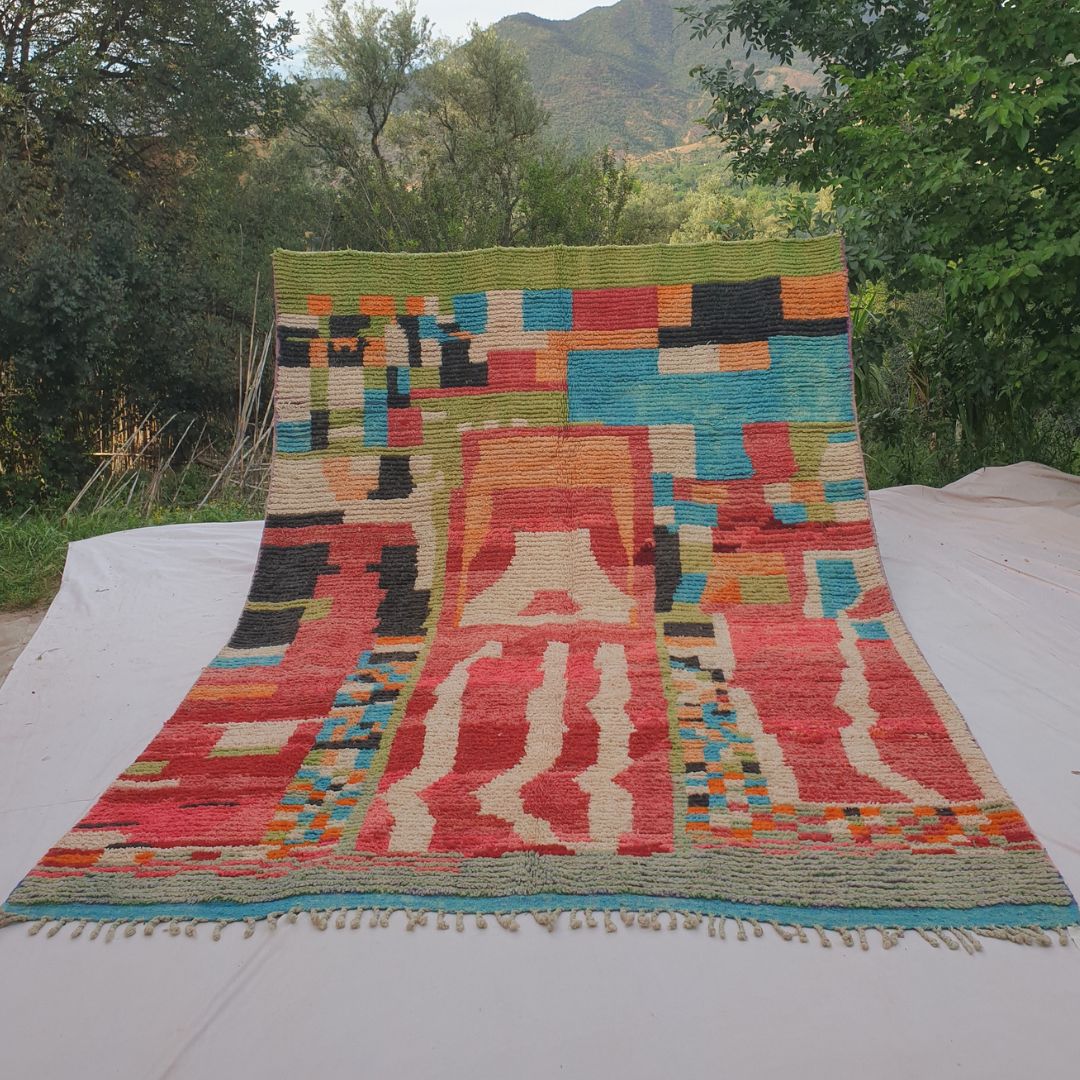 Person holding up a vibrant red multicolor Boujad Moroccan rug outdoors between trees, showing its large 254×349 cm size with colorful geometric patches in green, turquoise, orange, and black on a coral-red wool background
