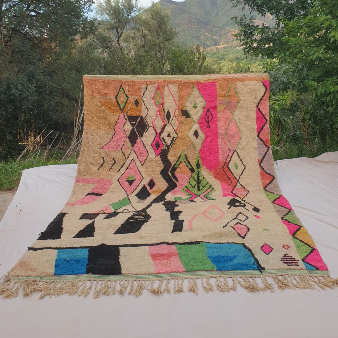 Person holding up a large cream Boujad Moroccan rug outdoors between trees, displaying its 259×359 cm size with bold black geometric shapes, fuchsia pink diamond outlines and green botanical motifs on an ivory wool background with a vibrant striped bottom band in green, blue and pink