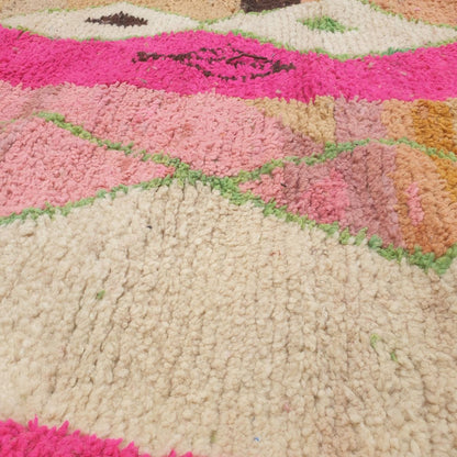 Close-up of a handwoven Boujad Berber rug showing bold graphic black geometric forms, intricate pink diamond outlines and green leaf motifs on a cream wool pile — a striking abstract design with clear Amazigh symbolic roots
