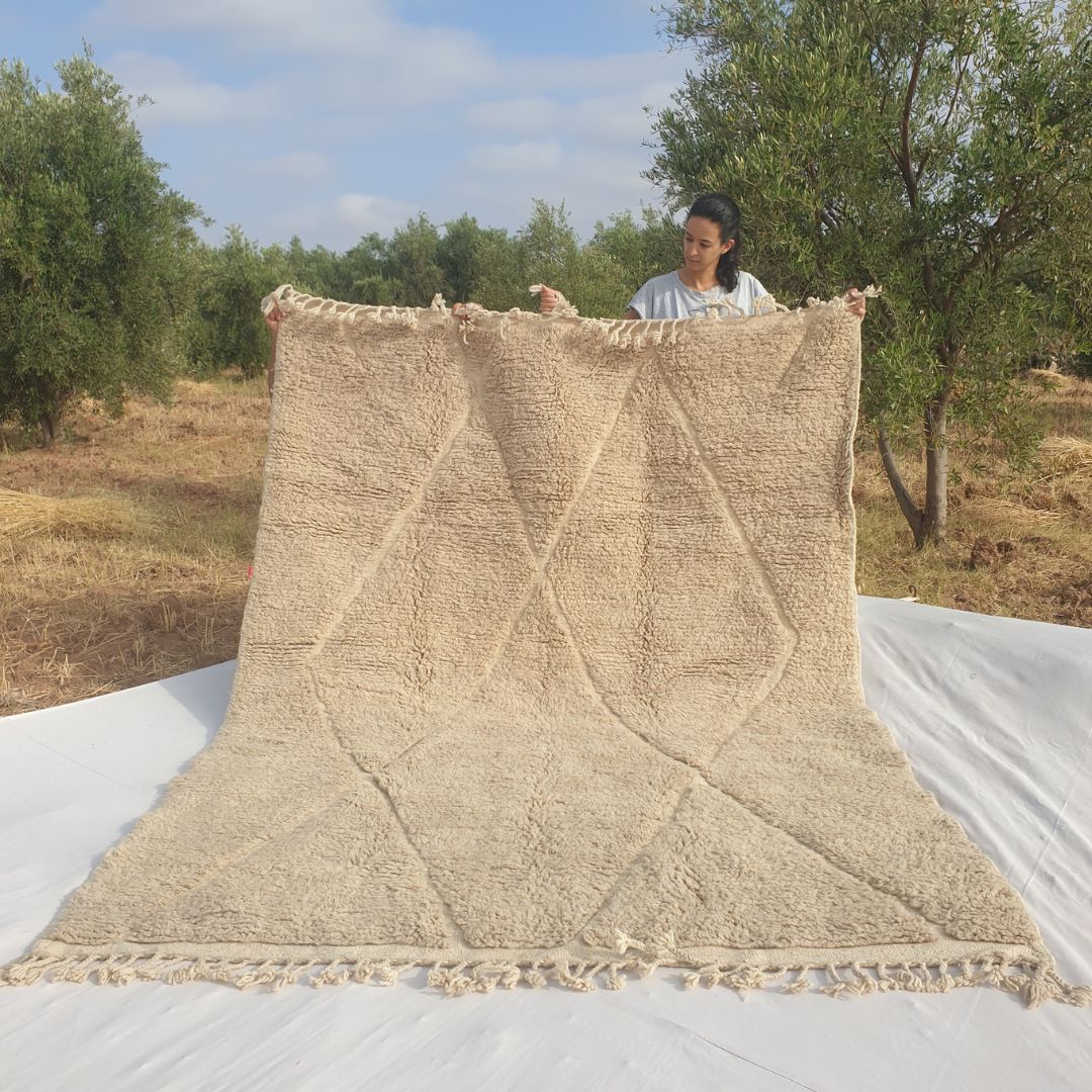 Person holding up an ivory beige Beni Ourain Moroccan rug outdoors, displaying its 207x281 cm size - a warm natural ivory and greige undyed wool base with a full-field classic diamond trellis pattern carved entirely through pile height variation, creating a tone-on-tone relief effect with natural fringe at both ends