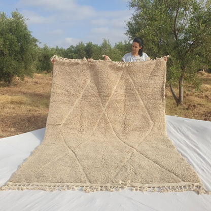 Person holding up an ivory beige Beni Ourain Moroccan rug outdoors, displaying its 207x281 cm size - a warm natural ivory and greige undyed wool base with a full-field classic diamond trellis pattern carved entirely through pile height variation, creating a tone-on-tone relief effect with natural fringe at both ends
