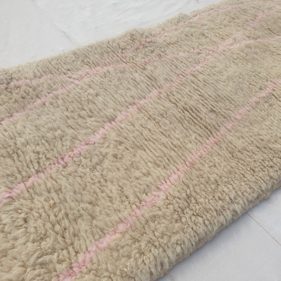 Close-up of a Cotya ivory Beni Ourain runner showing the soft natural wool pile with sparse dusty pink diagonal lines - the lines crisscrossing at wide intervals rather than forming a dense pattern, the restraint of the composition allowing the wool itself to remain the focus