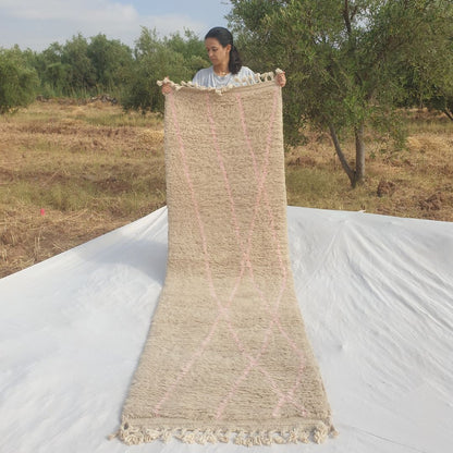 Person holding up an ivory Beni Ourain Moroccan runner rug outdoors, displaying its 84x284 cm size - a luminous natural ivory wool base with widely-spaced delicate dusty pink diagonal lines crossing the field sparingly, leaving large areas of plain ivory wool visible, with natural fringe at both ends