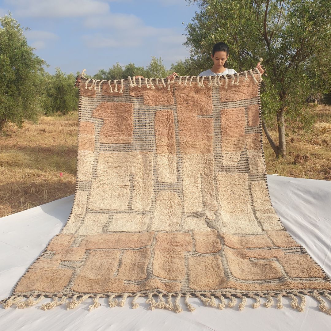 Person holding up a two-tone Beni Ourain Moroccan rug outdoors, displaying its 196x280 cm size - large irregular blocks of soft blush-rose and natural ivory/white wool arranged in an organic patchwork composition, separated by thin grey-blue grid lines throughout, with knotted tasselled fringe at both ends