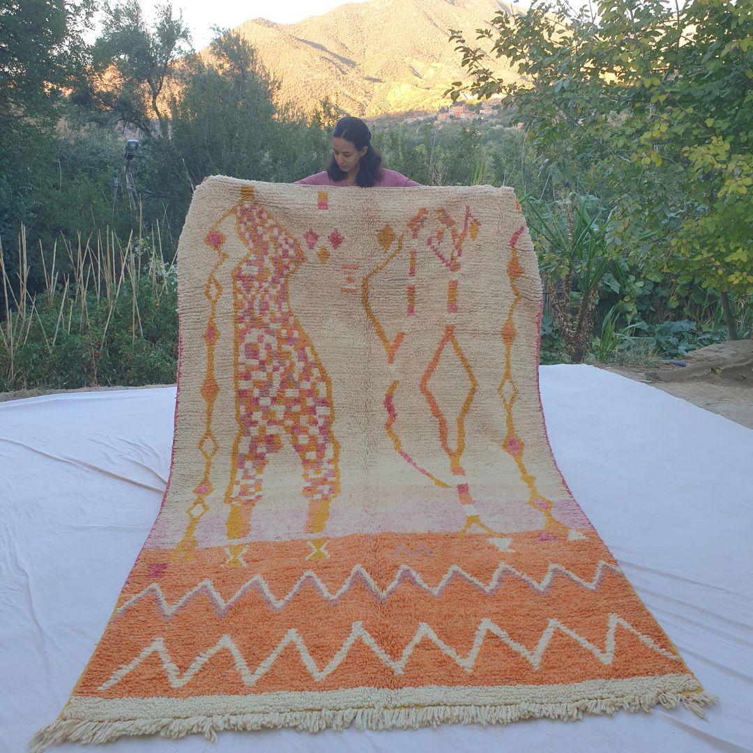 Large 6x10 orange and beige Boujad Moroccan rug featuring rare abstract tribal human or animal figures, diamonds, zigzags and symbolic motifs, displayed outdoors at sunset with mountain view