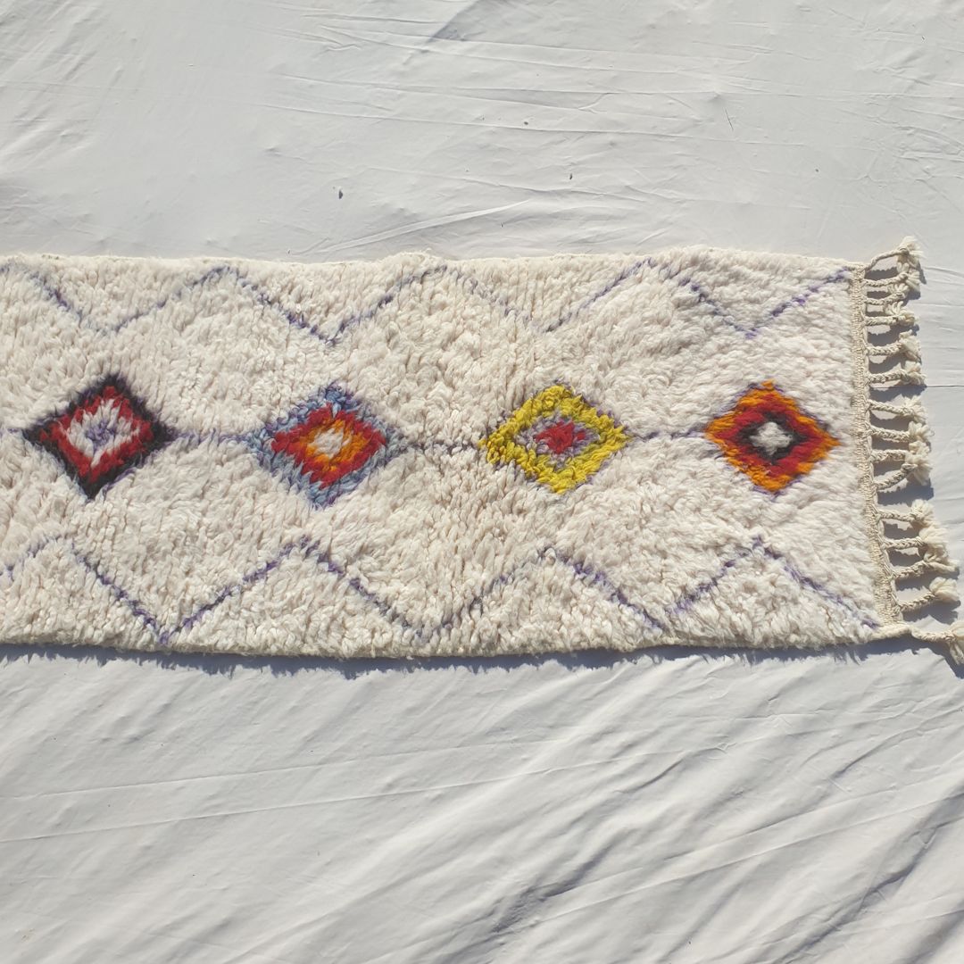 Bottom half of a white Beni Ourain Moroccan runner showing the fine diamond lattice background across the ivory wool pile and the lower solid-filled multicolor diamonds of the central column in warm orange, yellow and wine tones, with natural knotted fringe