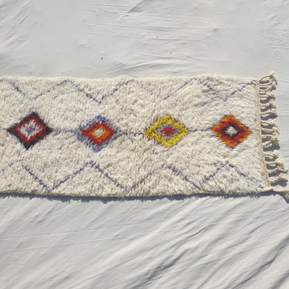 Bottom half of a white Beni Ourain Moroccan runner showing the fine diamond lattice background across the ivory wool pile and the lower solid-filled multicolor diamonds of the central column in warm orange, yellow and wine tones, with natural knotted fringe