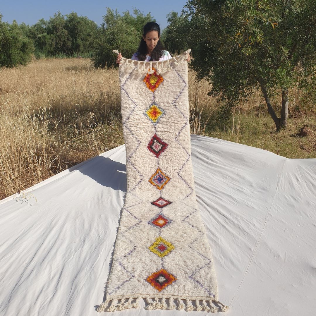 Person holding up a white Beni Ourain Moroccan runner rug outdoors, displaying its 69×290 cm size — an ivory natural wool base with a continuous fine diamond lattice pattern across the entire field and a central column of solid-filled multicolor diamonds in burnt orange, rust, golden yellow, wine red and warm mauve, each in a distinct warm earthy colour