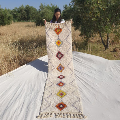 Person holding up a white Beni Ourain Moroccan runner rug outdoors, displaying its 69×290 cm size — an ivory natural wool base with a continuous fine diamond lattice pattern across the entire field and a central column of solid-filled multicolor diamonds in burnt orange, rust, golden yellow, wine red and warm mauve, each in a distinct warm earthy colour