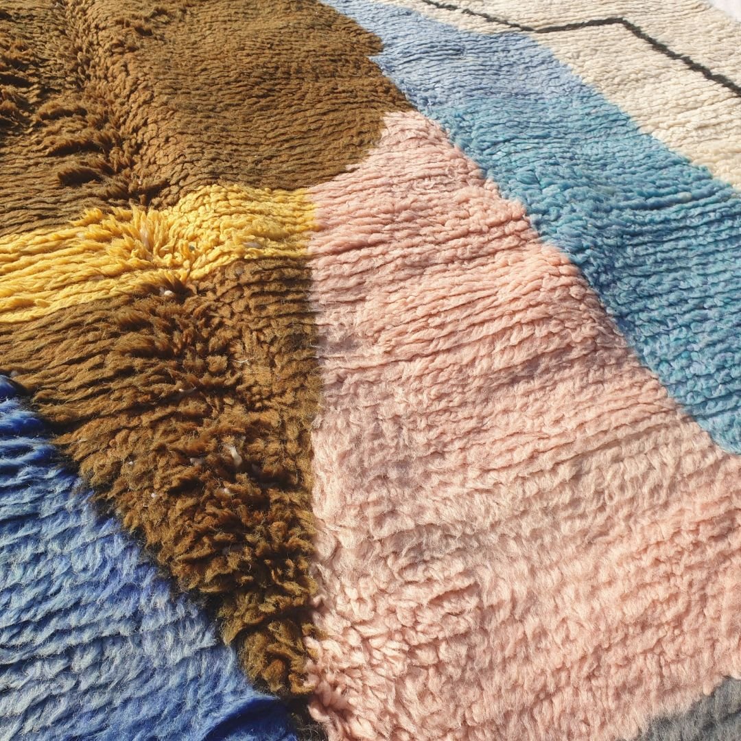 Detailed abstract geometric motifs including central diamond, triangles and color blocks in soft blue, pastel pink, warm beige, brown and yellow on authentic Beni Ourain shag rug