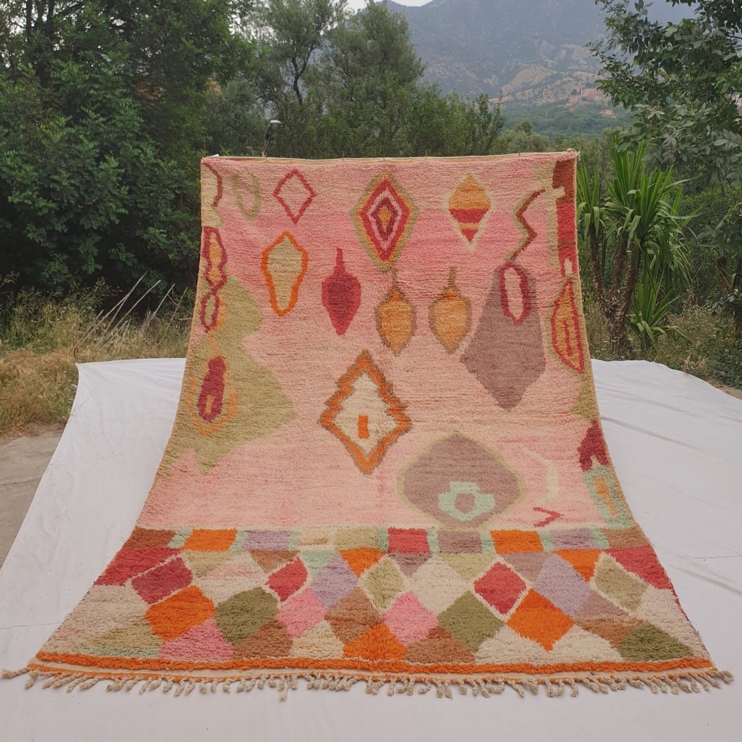 Person holding up a rose pink Moroccan rug outdoors, displaying its 200x303 cm size - a clear rose pink field scattered with pendant and leaf-shaped motifs in olive, red and orange, transitioning into a bright rainbow harlequin diamond band across the bottom third