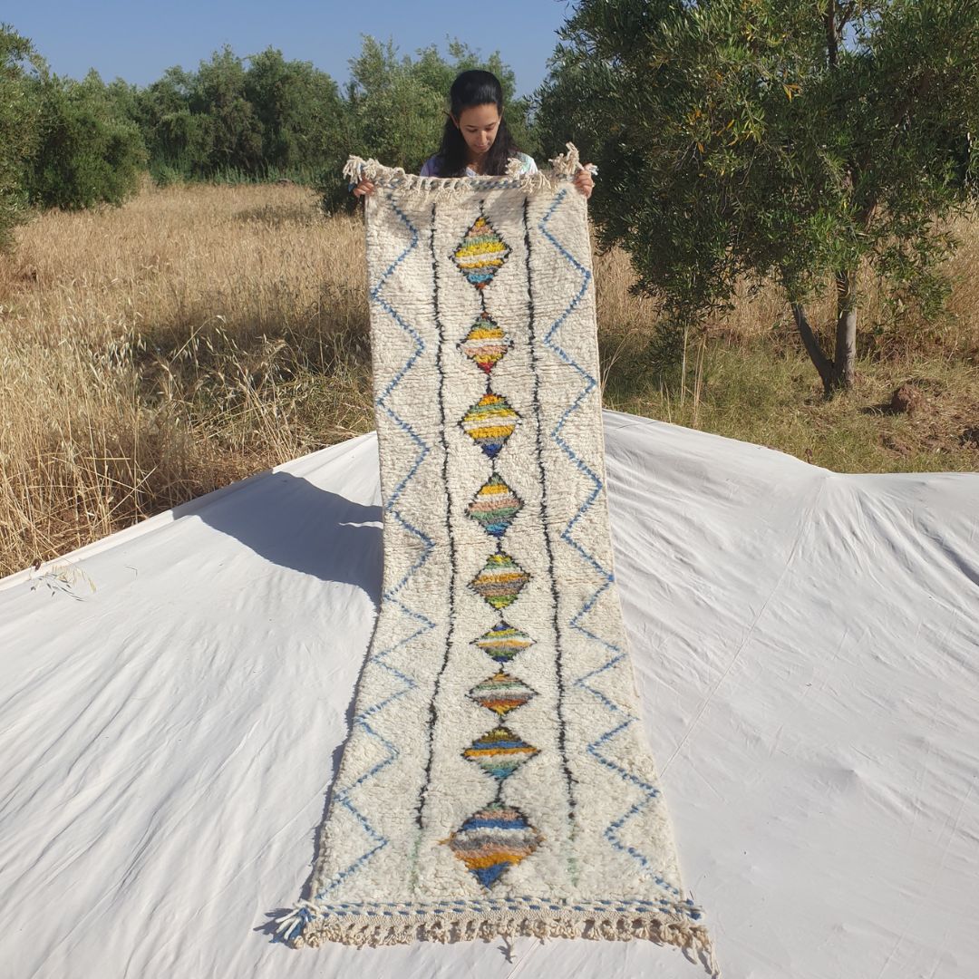 Person holding up a white Beni Ourain Moroccan runner rug outdoors, displaying its 75×282 cm size — an ivory natural wool base featuring a central column of multicolor striped diamonds in yellow, orange, teal, green, red, pink and grey running the full length, flanked by parallel black lines and a delicate blue zigzag chevron border