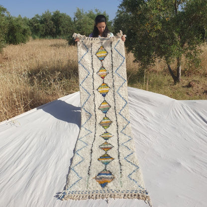 Person holding up a white Beni Ourain Moroccan runner rug outdoors, displaying its 75×282 cm size — an ivory natural wool base featuring a central column of multicolor striped diamonds in yellow, orange, teal, green, red, pink and grey running the full length, flanked by parallel black lines and a delicate blue zigzag chevron border