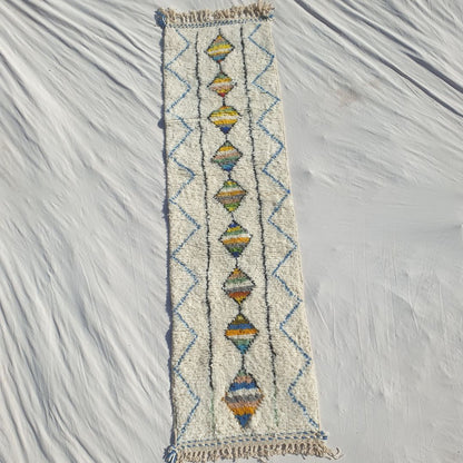 Authentic Beni Ourain Moroccan runner rug 75×282 cm laid flat — white cream natural wool base with a central column of multicolor striped diamonds running its full length, each diamond individually coloured in unique stripe combinations, parallel black flanking lines and a blue zigzag border, handwoven in Morocco