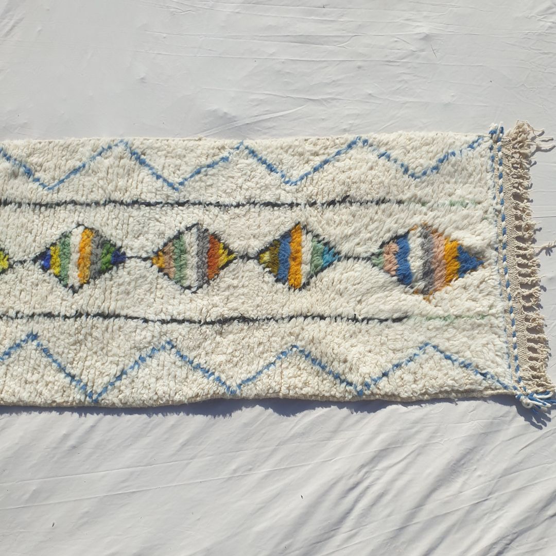 Bottom half of a white Beni Ourain Moroccan runner showing the natural ivory wool pile with the final multicolor striped diamonds of the central column, the flanking black vertical lines and the delicate blue chevron outer border, with natural knotted fringe — the exceptional softness of authentic Beni Ourain High Atlas wool
