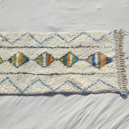 Bottom half of a white Beni Ourain Moroccan runner showing the natural ivory wool pile with the final multicolor striped diamonds of the central column, the flanking black vertical lines and the delicate blue chevron outer border, with natural knotted fringe — the exceptional softness of authentic Beni Ourain High Atlas wool