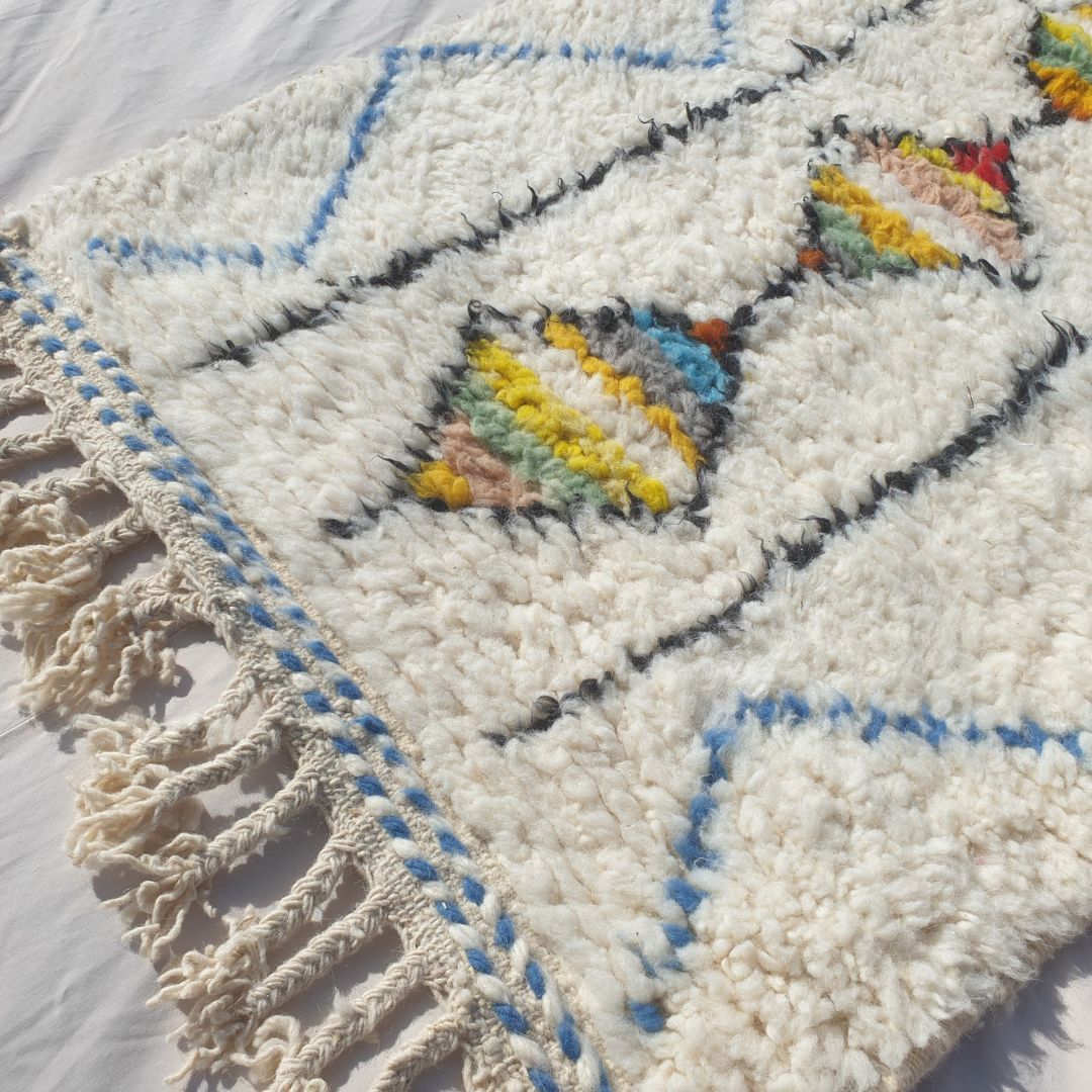 Close-up of a white Beni Ourain runner rug showing the extraordinary natural ivory wool pile with a central column of striped diamond motifs — each diamond filled with unique horizontal colour bands in yellow, orange, teal, green, red and pink, framed by parallel black lines and a blue zigzag outer border, all handwoven in 100% natural wool