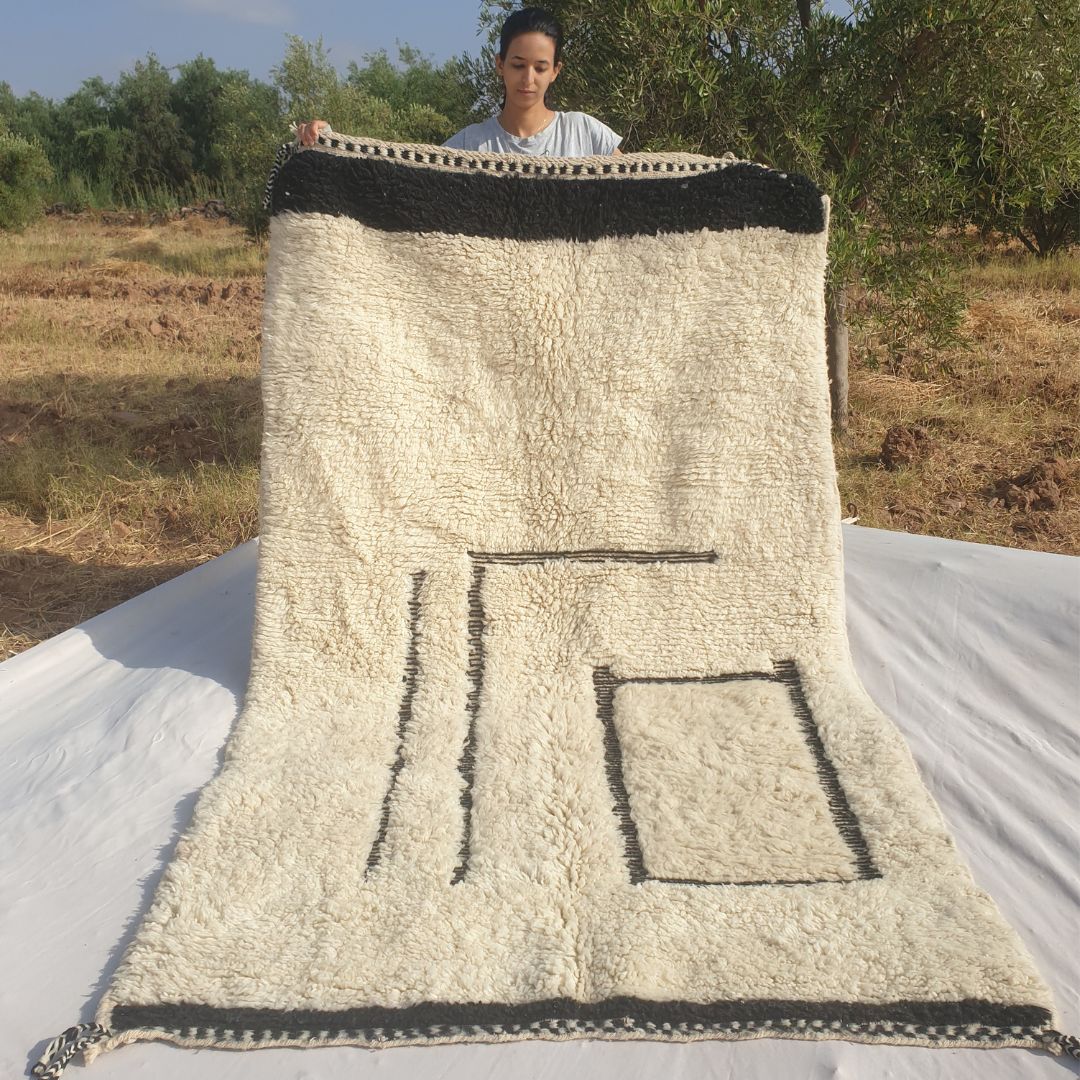 Person holding up a black and white Beni Ourain Moroccan rug outdoors, displaying its 154x264 cm size - a clean ivory white wool base with an asymmetric minimalist black line composition: two bold vertical parallel lines on the left side and a standalone black rectangle outline in the lower right area, with a solid black border stripe at top and bottom