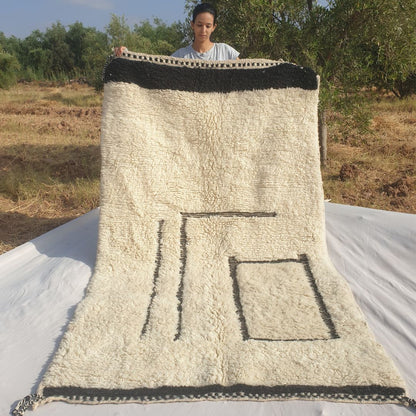 Person holding up a black and white Beni Ourain Moroccan rug outdoors, displaying its 154x264 cm size - a clean ivory white wool base with an asymmetric minimalist black line composition: two bold vertical parallel lines on the left side and a standalone black rectangle outline in the lower right area, with a solid black border stripe at top and bottom