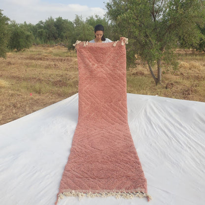 Person holding up a terracotta rose pink Beni Ourain Moroccan runner rug outdoors, displaying its 88x325 cm size - a single saturated dusty rose terracotta wool field with a carved diagonal diamond relief pattern visible only through subtle pile height variation, with natural fringe at both ends