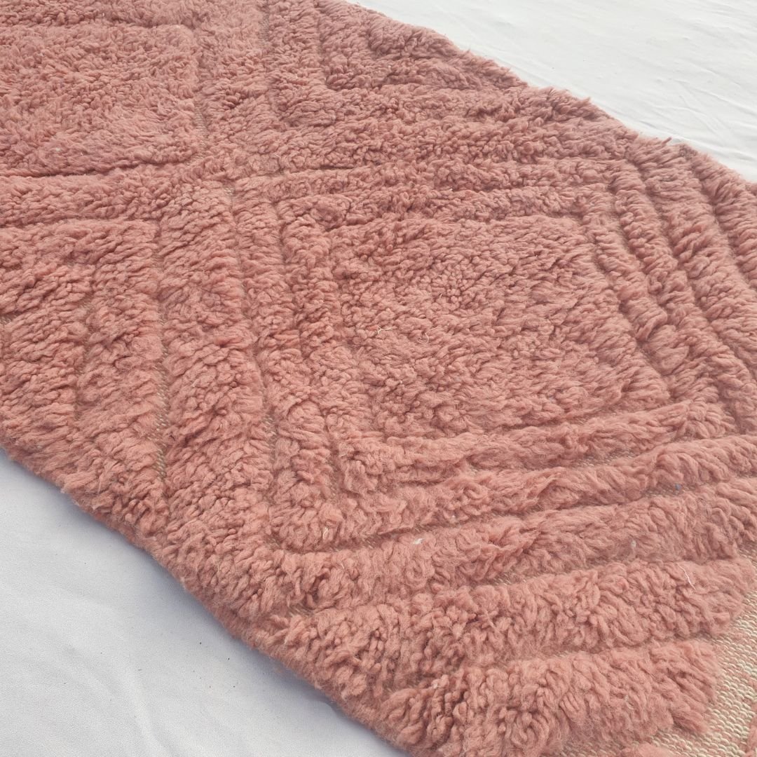 Close-up of a Karisa terracotta rose Beni Ourain runner showing the rich saturated pink wool pile with a carved diagonal diamond relief texture - the diamonds visible only through shifts in light across the pile height, an entirely tonal monochromatic composition handwoven in 100% natural wool