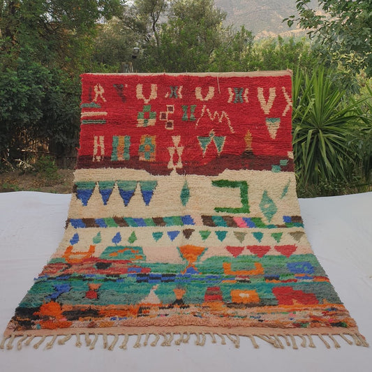 Vibrant 6x9 Boujad Moroccan rug featuring bold tribal symbols, diamonds, arrows, geometric shapes and abstract motifs in rich red, orange, pink, green, blue and black on creamy ivory wool, high-pile texture, displayed outdoors with mountain view