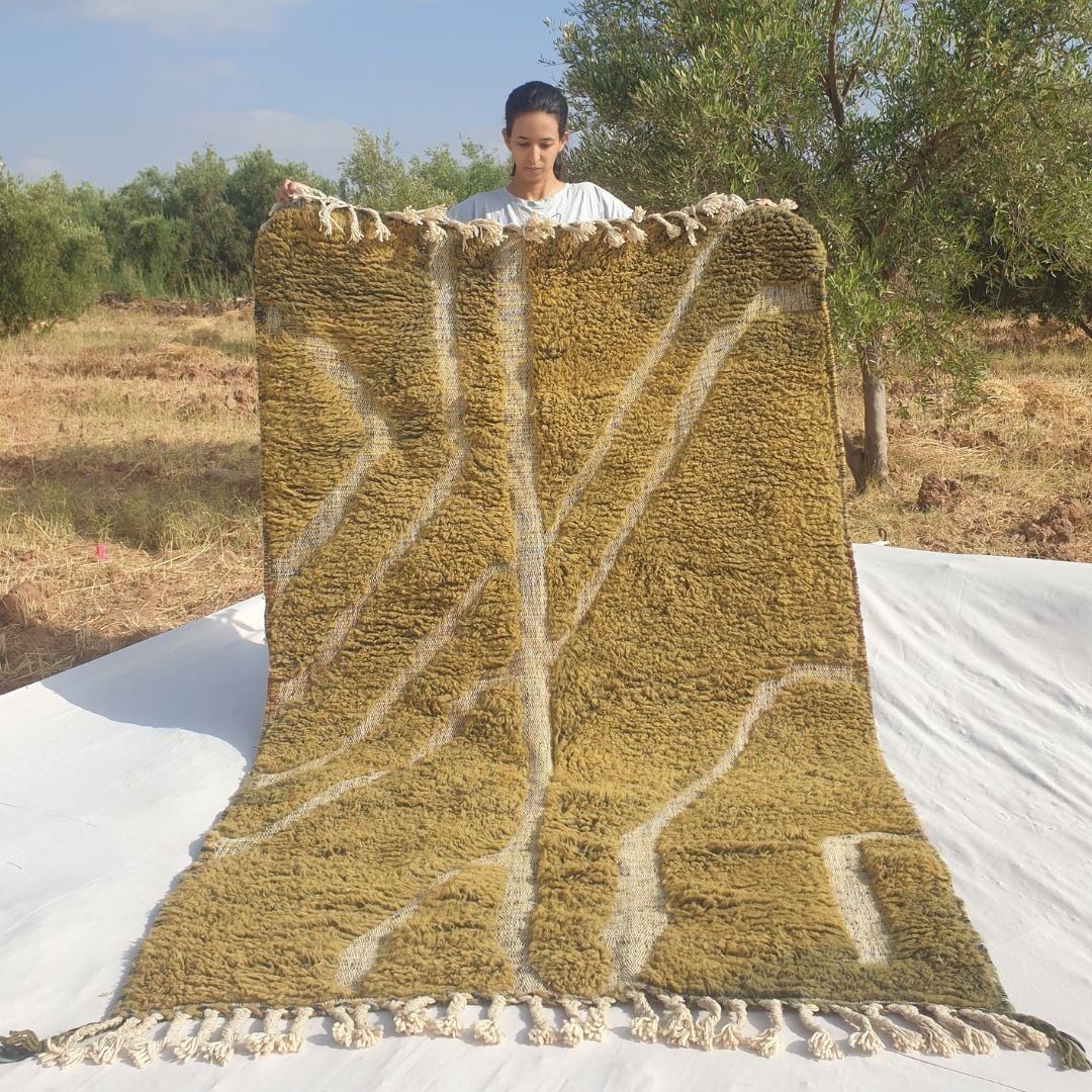 Person holding up an olive green Beni Ourain Moroccan rug outdoors, displaying its 153x242 cm size - a rich warm olive and moss green wool base with an abstract organic branching ivory line composition: a central stem-like line branching into sweeping diagonal curves left and right, with a rectangular accent in the lower right, natural fringe at both ends