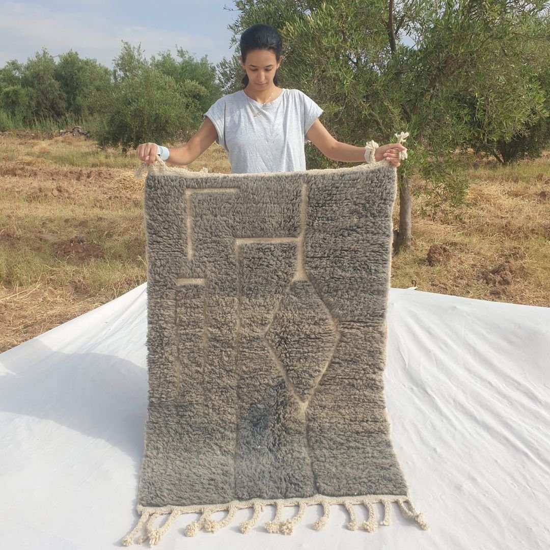 Person holding up a grey taupe Beni Ourain Moroccan accent rug outdoors, displaying its 100x145 cm size - a muted smoky grey taupe wool base with an asymmetric dark line composition: an irregular stepped frame of vertical lines on the left and a diamond shape formed where two angled lines meet on the right, with natural fringe at both ends