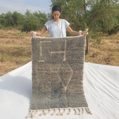 Person holding up a grey taupe Beni Ourain Moroccan accent rug outdoors, displaying its 100x145 cm size - a muted smoky grey taupe wool base with an asymmetric dark line composition: an irregular stepped frame of vertical lines on the left and a diamond shape formed where two angled lines meet on the right, with natural fringe at both ends