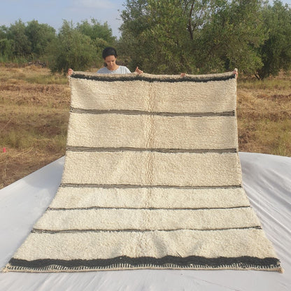 Person holding up an ivory Beni Ourain Moroccan rug outdoors, displaying its 211x284 cm size - a natural undyed wool base with clusters of black horizontal stripes of varying thickness running across the width, and a black-and-white checkered woven border at one end, with natural fringe