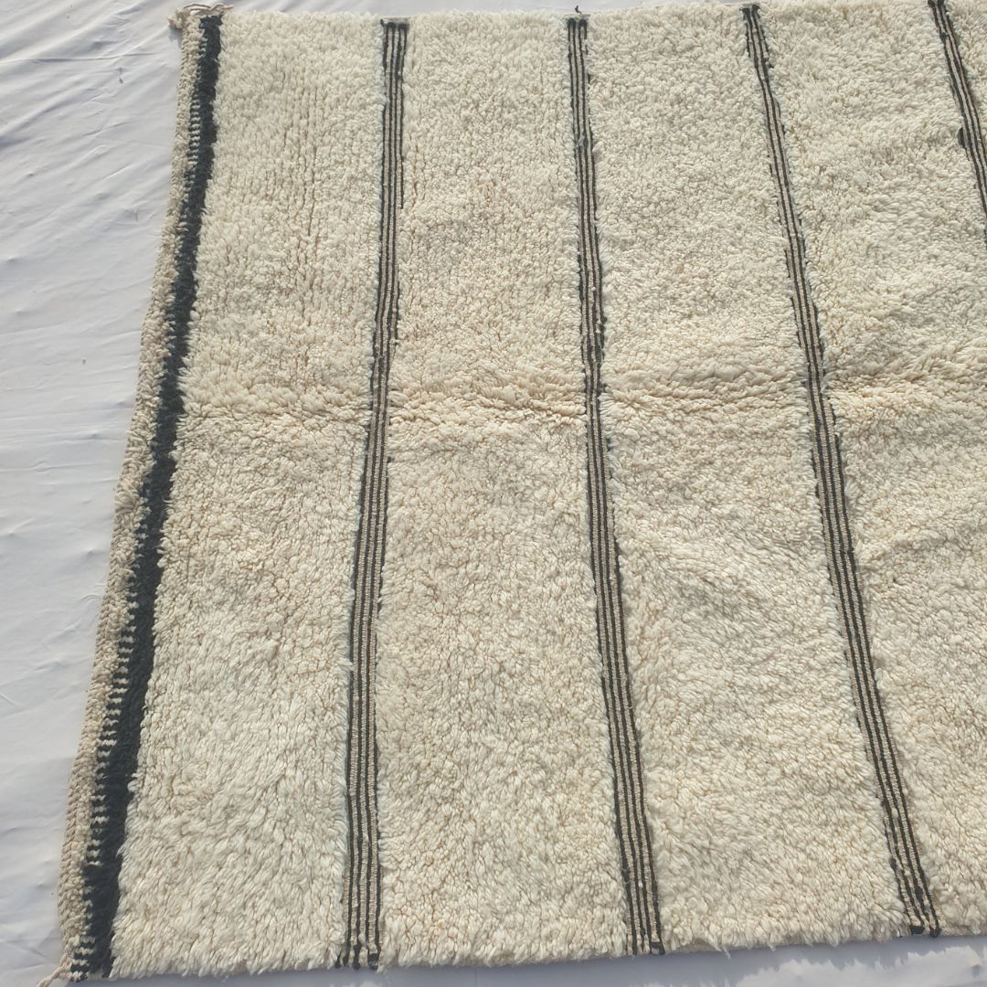 Bottom half of an ivory Beni Ourain Moroccan rug showing the black-and-white checkered border and the final black stripe clusters against the natural wool pile, with hand-knotted fringe - the exceptional softness of authentic Beni Ourain wool