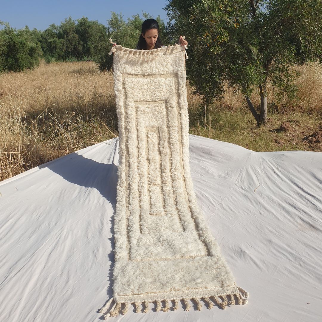 Person holding up a pure white Beni Ourain Moroccan runner rug outdoors, displaying its 74×290 cm size — a natural undyed ivory wool runner featuring a subtle carved relief composition of nested concentric rectangles creating a frame-within-frame architectural pattern through pile height variation, with natural fringe at both ends