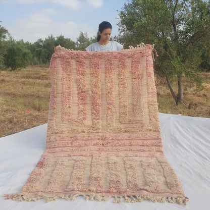 Person holding up a dusty rose pink Beni Ourain Moroccan rug outdoors, displaying its 148x237 cm size - a warm muted blush pink wool base with a tonal darker rose geometric maze pattern of two mirrored rectangular panels containing concentric U-shaped comb forms nesting within each other, with natural fringe at both ends