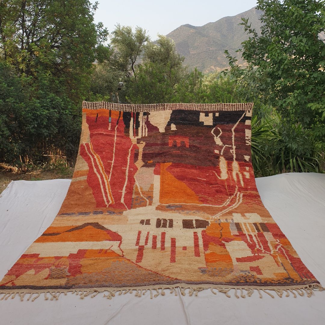 Large 9x12 Boujad Moroccan rug with vibrant abstract tribal composition in warm oranges, deep reds, pinks, yellows, blues, greens and black on creamy ivory wool, high-pile texture, displayed outdoors with mountain backdrop