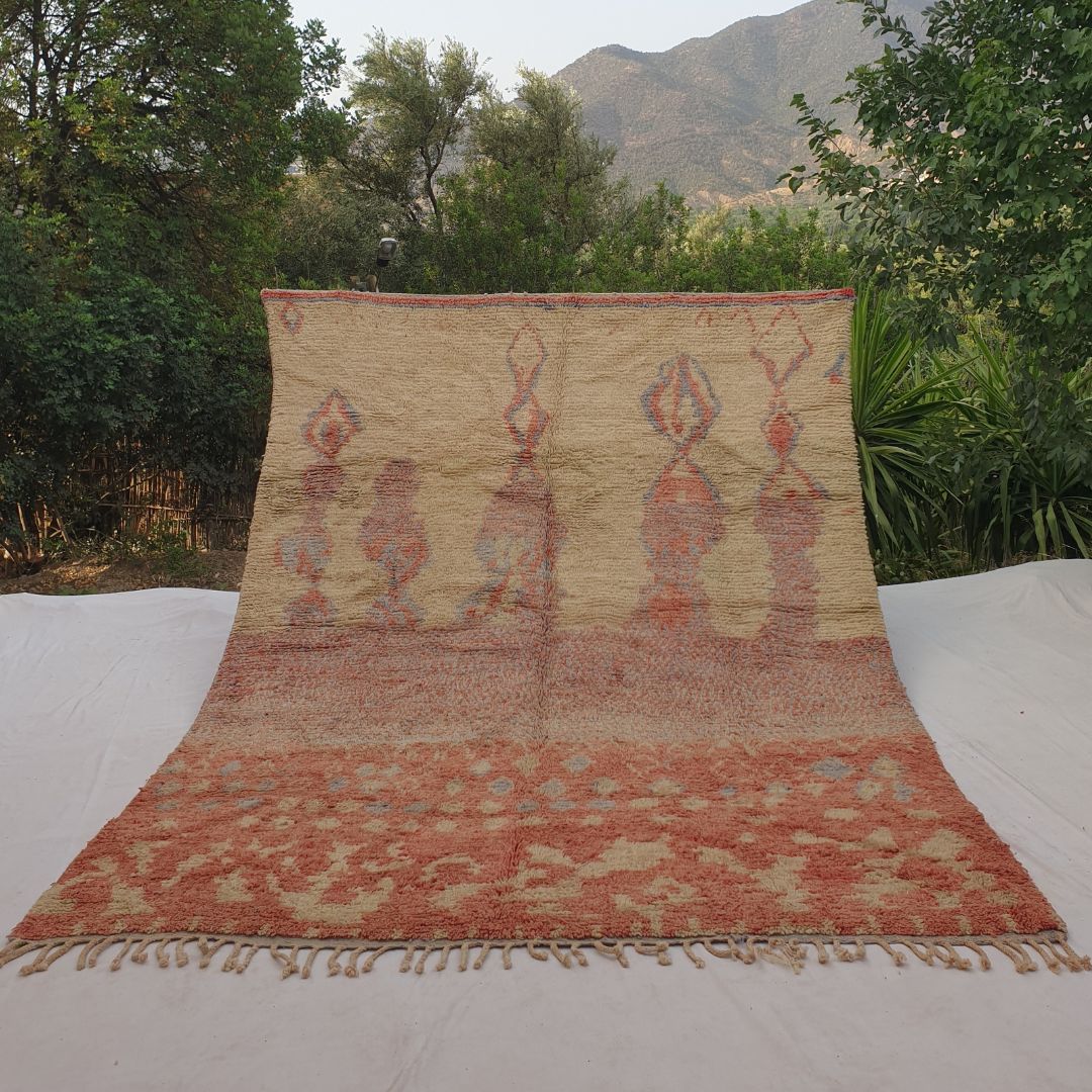 Large 8x11 Boujad Moroccan rug featuring vibrant abstract tribal motifs in soft peach, coral pink, warm orange, yellow, subtle blue and black on creamy ivory wool, high-pile texture, displayed outdoors with mountain backdrop