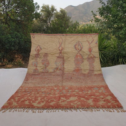 Large 8x11 Boujad Moroccan rug featuring vibrant abstract tribal motifs in soft peach, coral pink, warm orange, yellow, subtle blue and black on creamy ivory wool, high-pile texture, displayed outdoors with mountain backdrop