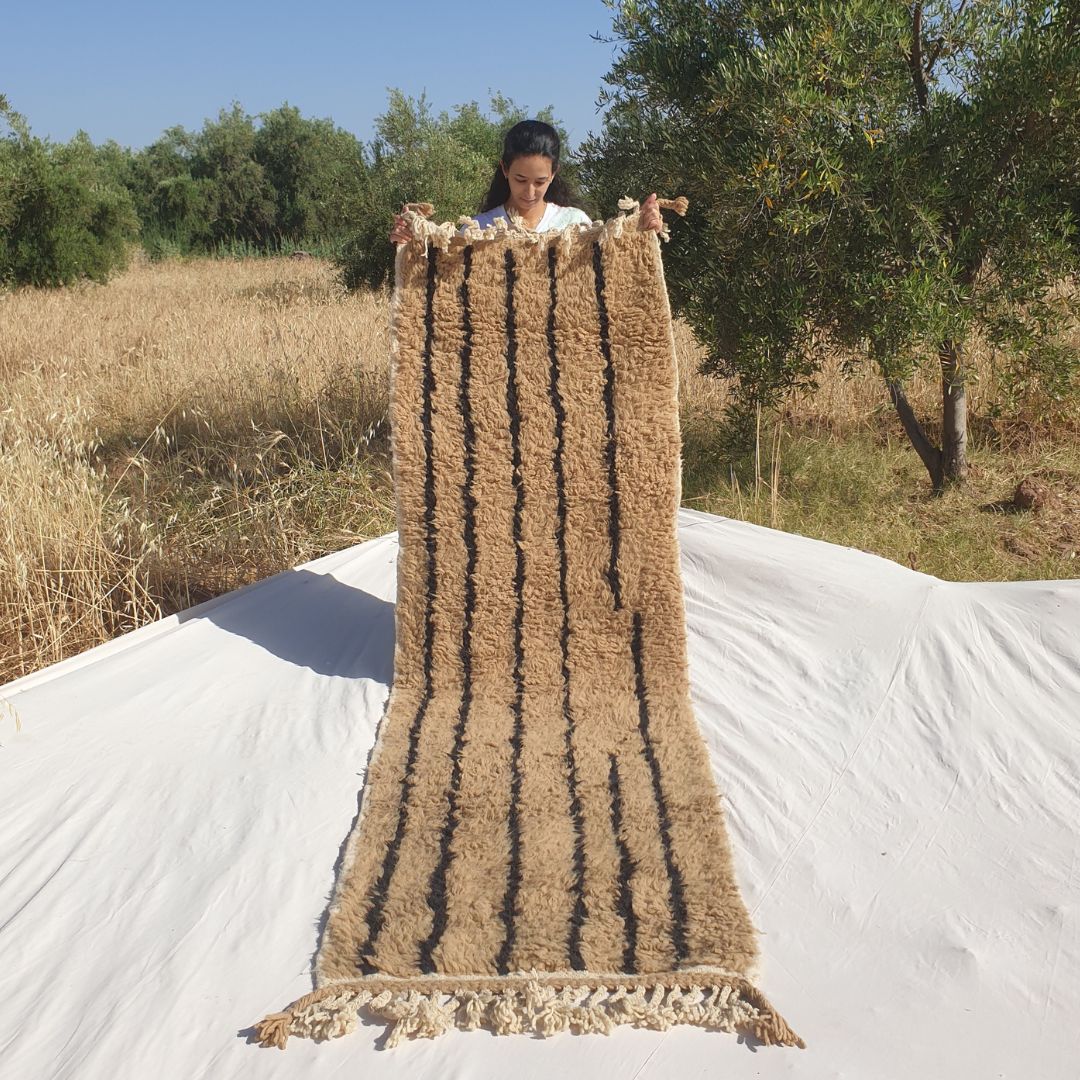 Person holding up a warm beige Beni Ourain Moroccan runner rug outdoors, displaying its 85x280 cm size - a natural camel and warm beige undyed wool base with clean vertical black stripe lines running the full length of the runner, creating a simple, architectural, deeply calming composition with natural fringe at both ends