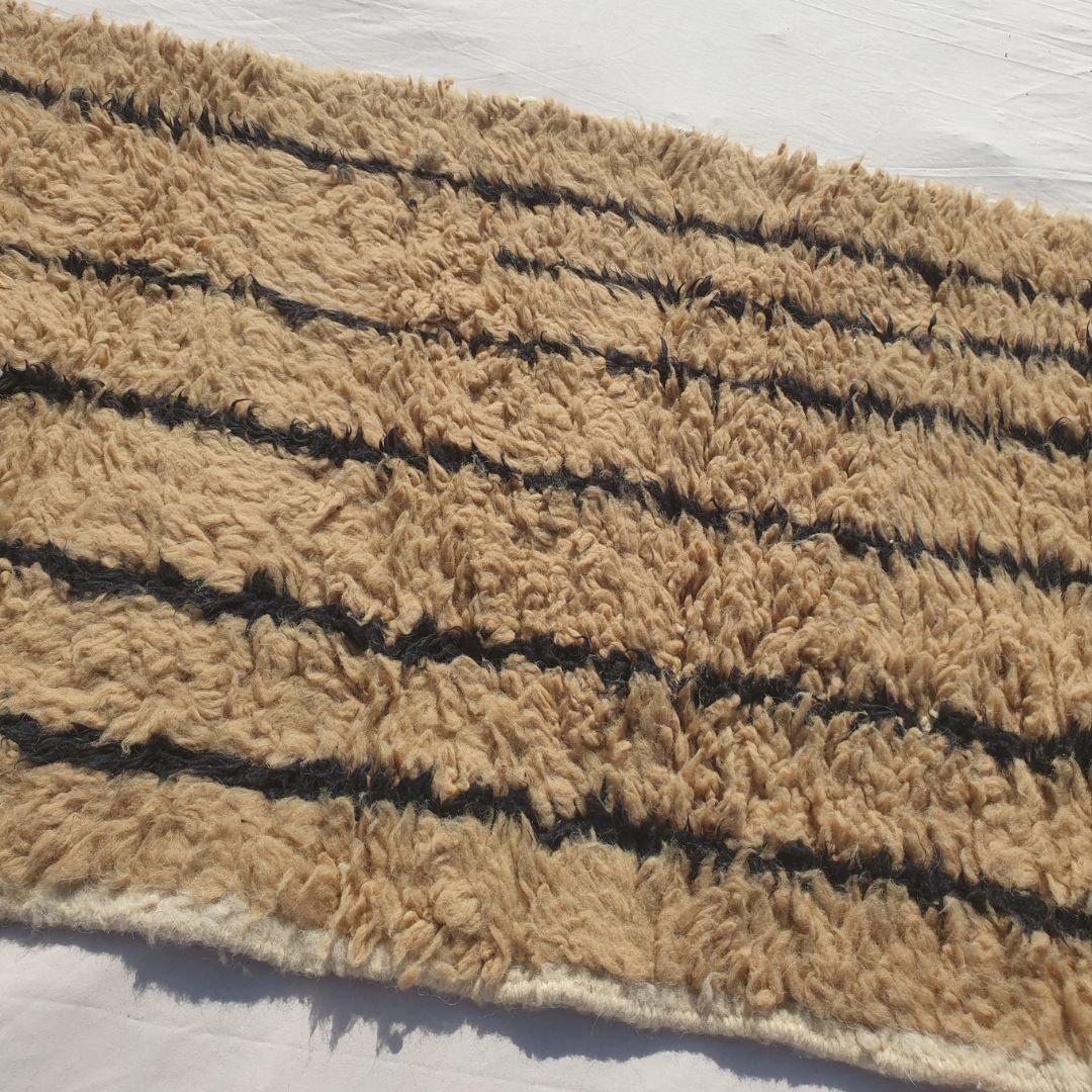 Close-up of a warm beige Beni Ourain runner rug showing the extraordinary softness of the natural camel and beige wool pile with precise vertical black stripe lines running along the full length - a minimalist two-tone composition of exceptional graphic clarity, all handwoven in 100% natural Atlas mountain wool