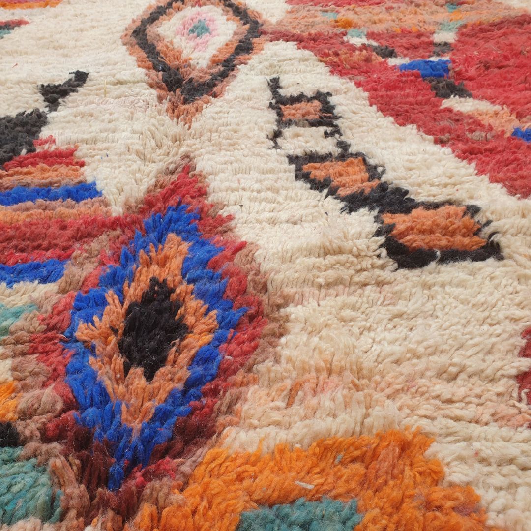 Close-up of a handwoven Boujad Berber rug showing intricate diamond medallion details in teal, pink, orange and black on a cream wool pile, surrounded by rich abstract multicolor geometric patches in blue, red and brown
