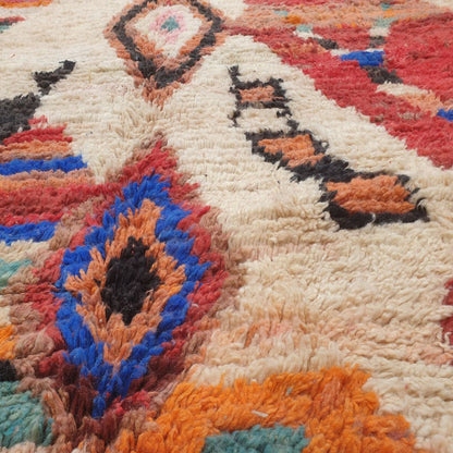 Close-up of a handwoven Boujad Berber rug showing intricate diamond medallion details in teal, pink, orange and black on a cream wool pile, surrounded by rich abstract multicolor geometric patches in blue, red and brown
