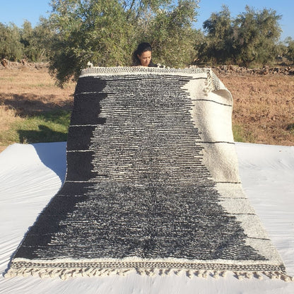 Classic black and white 6x10 Beni Ourain Moroccan rug featuring bold abstract geometric lines and stripes on creamy ivory background, ultra-plush high-pile texture, held outdoors in olive grove at sunset