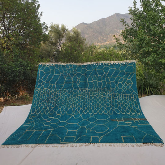 Rare oversized 10x10 square Beni Ourain Moroccan rug featuring intricate honeycomb geometric pattern in rich teal with creamy ivory outlines, ultra-fluffy high-pile texture, laid out outdoors against mountain backdrop and olive trees