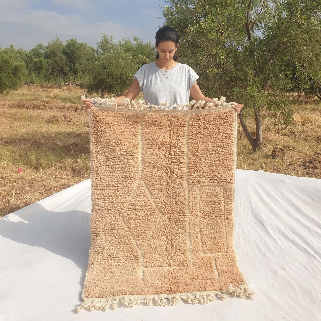 Person holding up a beige Beni Ourain Moroccan accent rug outdoors, displaying its 104x144 cm size - a warm golden beige wool base with a carved relief composition of a central diamond flanked by nested rectangular panels, entirely tone-on-tone with no colour contrast, natural fringe at both ends