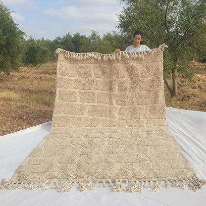 Person holding up an ivory Beni Ourain Moroccan rug outdoors, displaying its 201x314 cm size - a warm natural ivory undyed wool base with a full-field brick and cobblestone carved relief pattern, rows of irregular rectangular blocks separated by thin sunken lines both horizontally and vertically, entirely tone-on-tone, with natural fringe at both ends